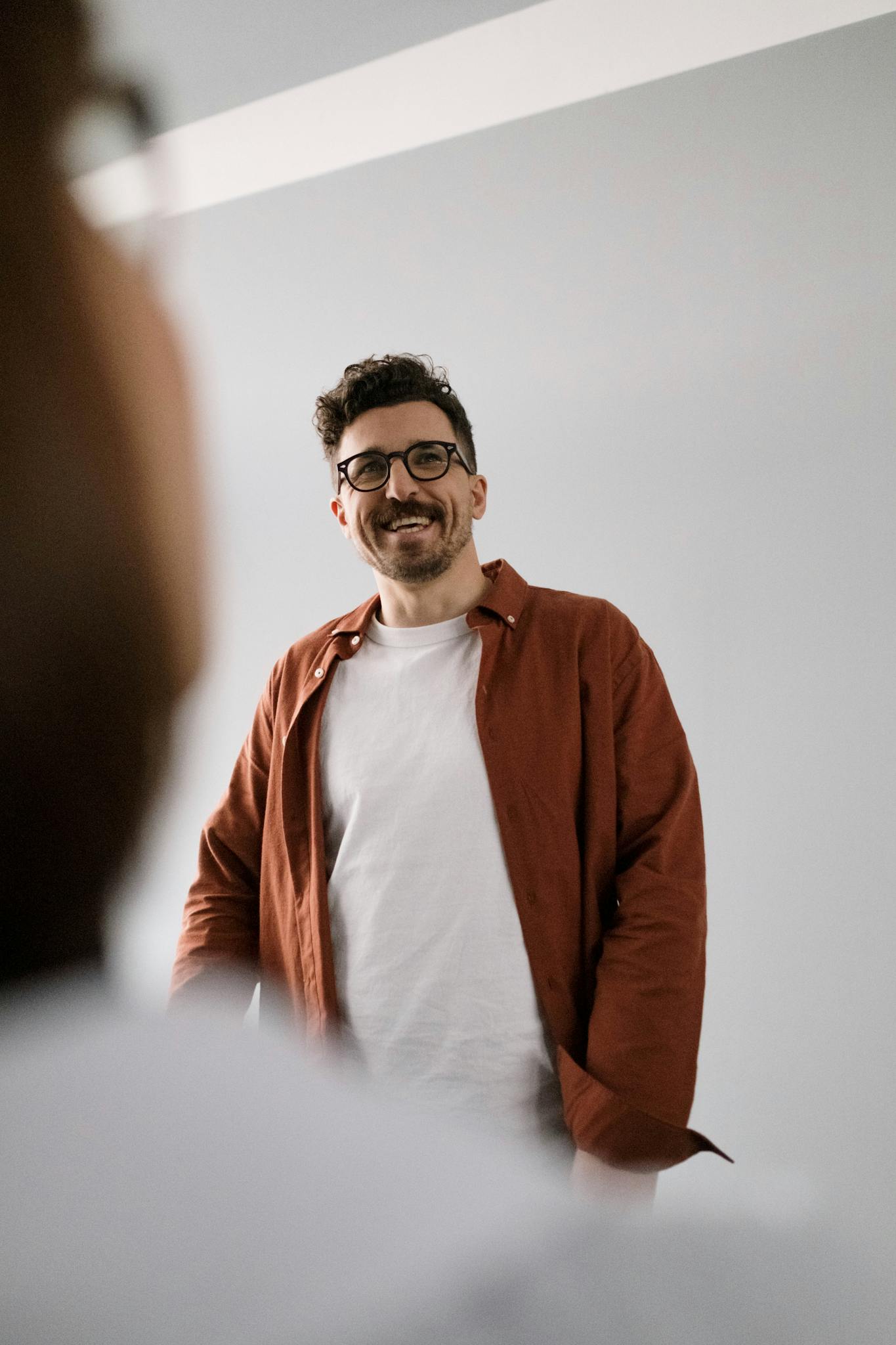 A cheerful man wearing glasses and a casual shirt standing indoors, captured naturally.
